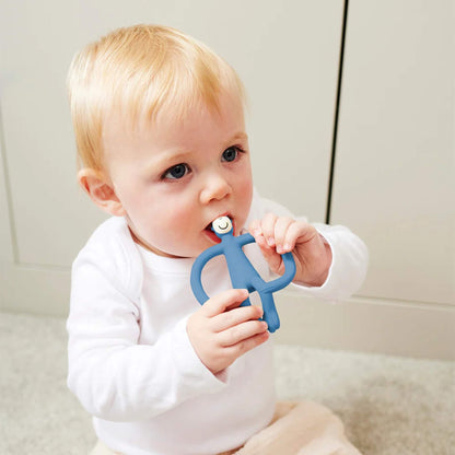 A baby sitting on the floor holding a blue silicone monkey teething Toy and biting the textured head while gripping the wide looped arms.