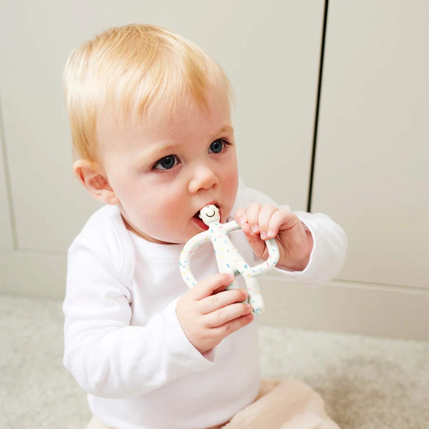A baby sitting on the floor chewing on a speckled silicone teether while holding it with both hands, wearing a white long-sleeve top and light-coloured trousers.