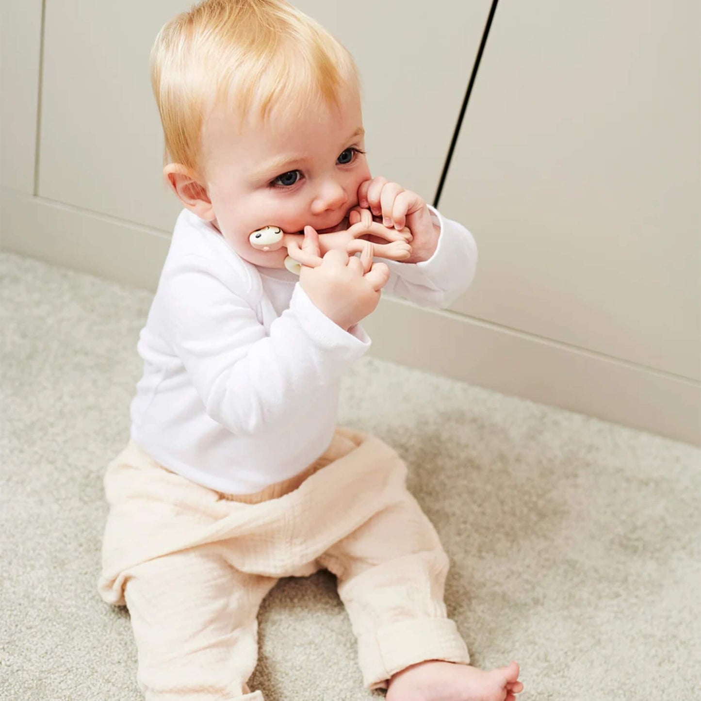 A baby with light blond hair sits on a soft carpet, gripping a soft pink silicone teether with both hands and chewing on one side. The child wears a white long-sleeved top and pale trousers with one leg slightly rolled up.