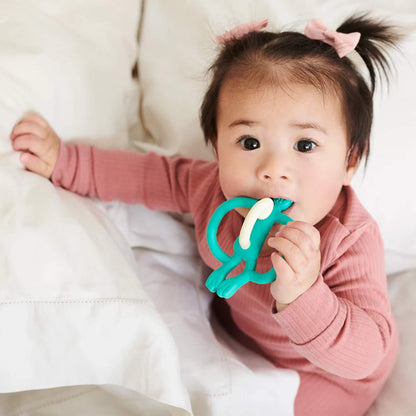 A baby sitting on a white bed holding a green silicone teether with both hands and chewing on the textured top section. The baby is wearing a long-sleeved pink ribbed outfit and has small bows in their hair.