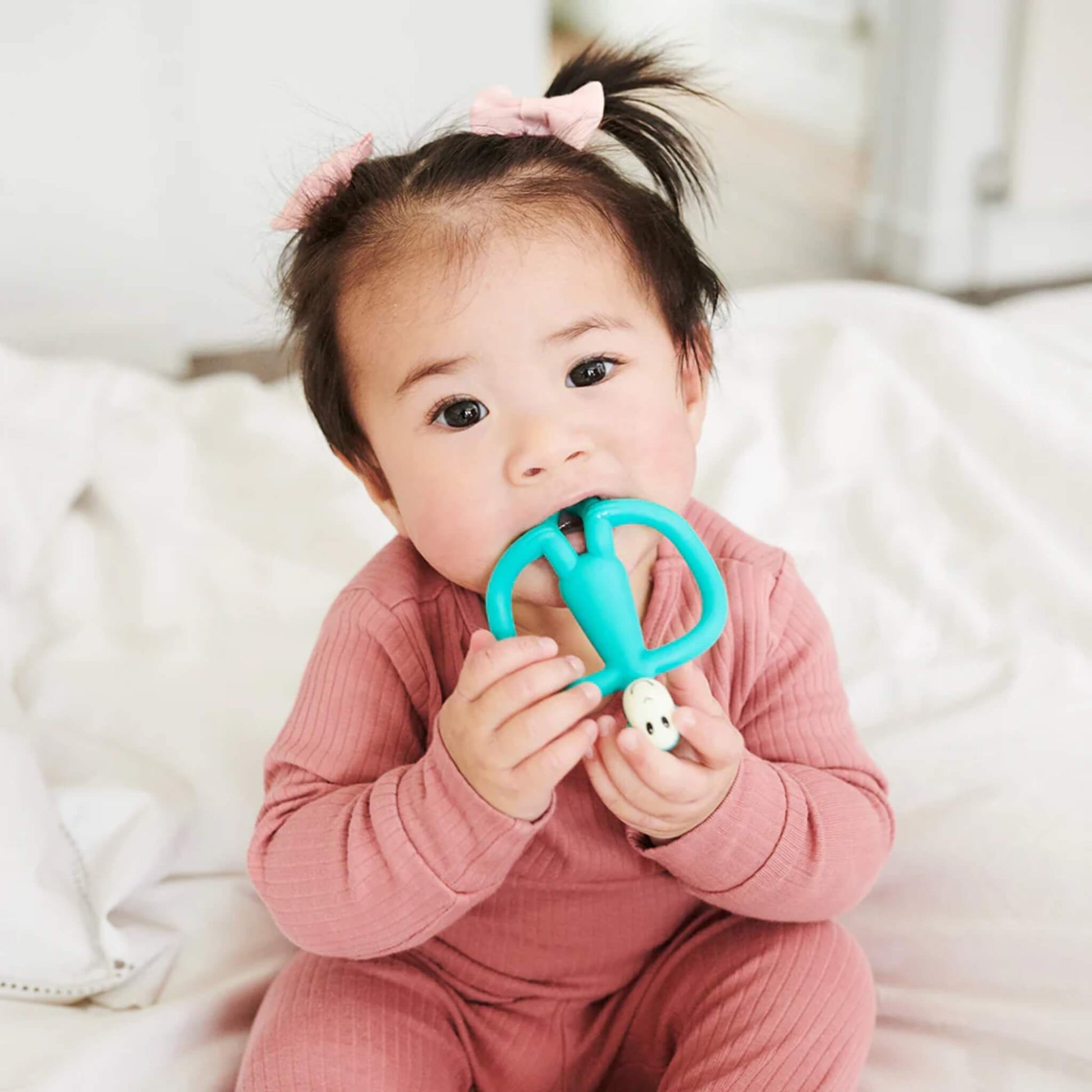 A baby sitting upright on a bed and chewing on a turquoise silicone teether while gripping it with both hands. The baby is dressed in a pink ribbed one-piece and has short pigtails with matching bows.