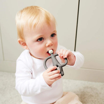 A baby sitting on a light-coloured floor holding a grey silicone teether with both hands and chewing on the smooth top section. The baby is wearing a white long-sleeved top and pale trousers.