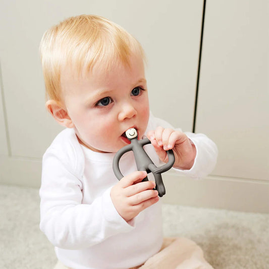 A baby sitting on a light-coloured floor holding a grey silicone teether with both hands and chewing on the smooth top section. The baby is wearing a white long-sleeved top and pale trousers.