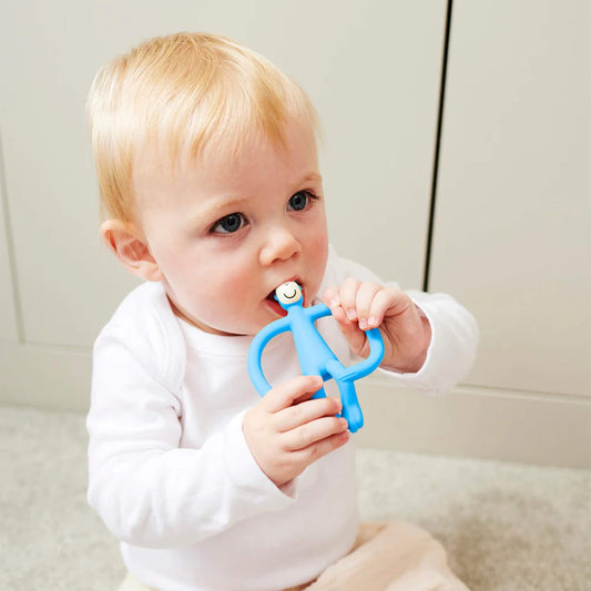 A baby sitting on a light-coloured floor holding a blue silicone teether with both hands and chewing on the smooth top section. The baby is wearing a white long-sleeved top and pale trousers.