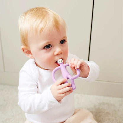 A baby sitting on a soft carpet holding a lilac monkey-shaped teether and chewing on the textured head.