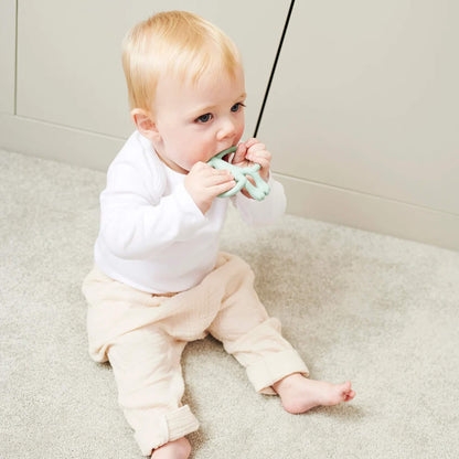 A baby sitting on a light-coloured floor holding a mint green silicone teether with both hands and chewing on the smooth top section. The baby is wearing a white long-sleeved top and pale trousers.