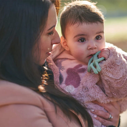 A baby being held outdoors while chewing on a mint-coloured silicone teether, wrapped in a soft pink coat with animal shapes on the front.
