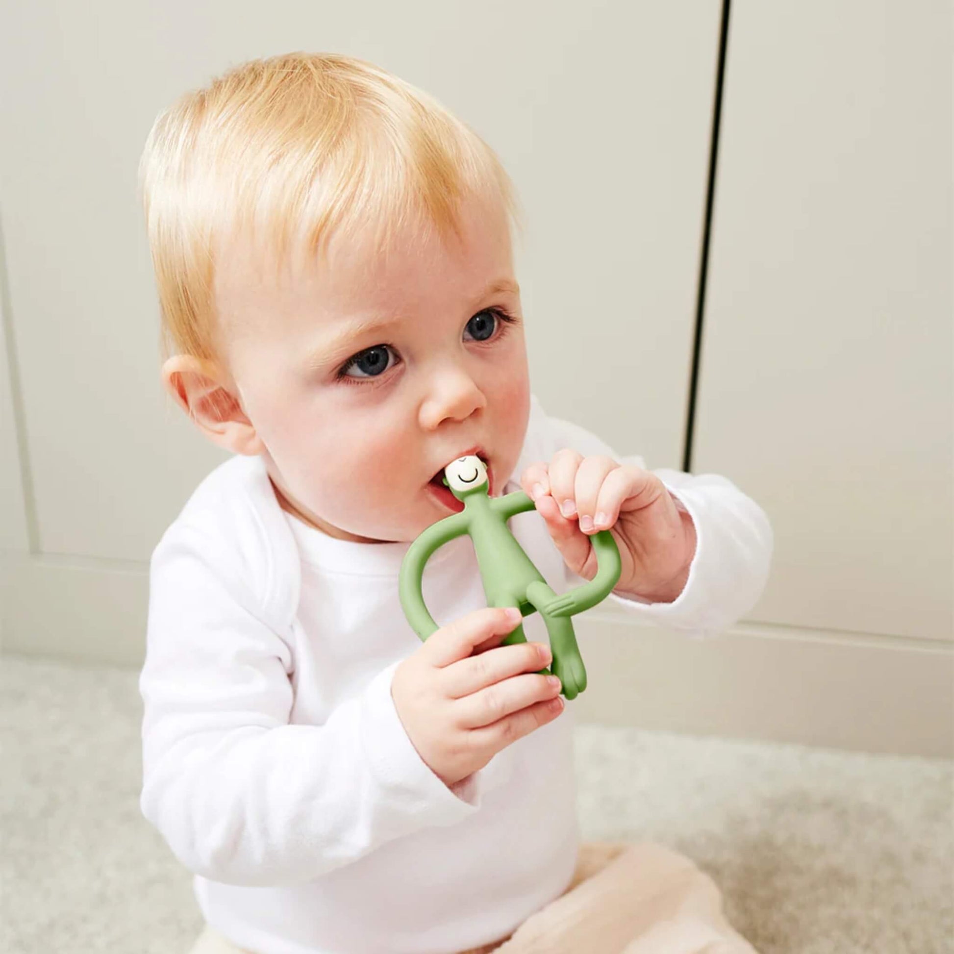 A baby sitting on a soft carpet holding an olive monkey-shaped teether and chewing on the textured head.
