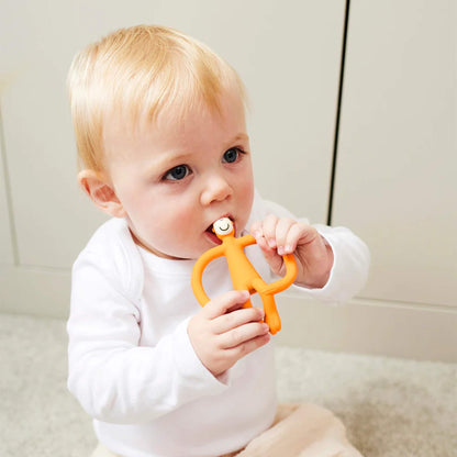 A baby sitting on a light-coloured floor holding an orange silicone teether with both hands and chewing on the smooth top section. The baby is wearing a white long-sleeved top and pale trousers.
