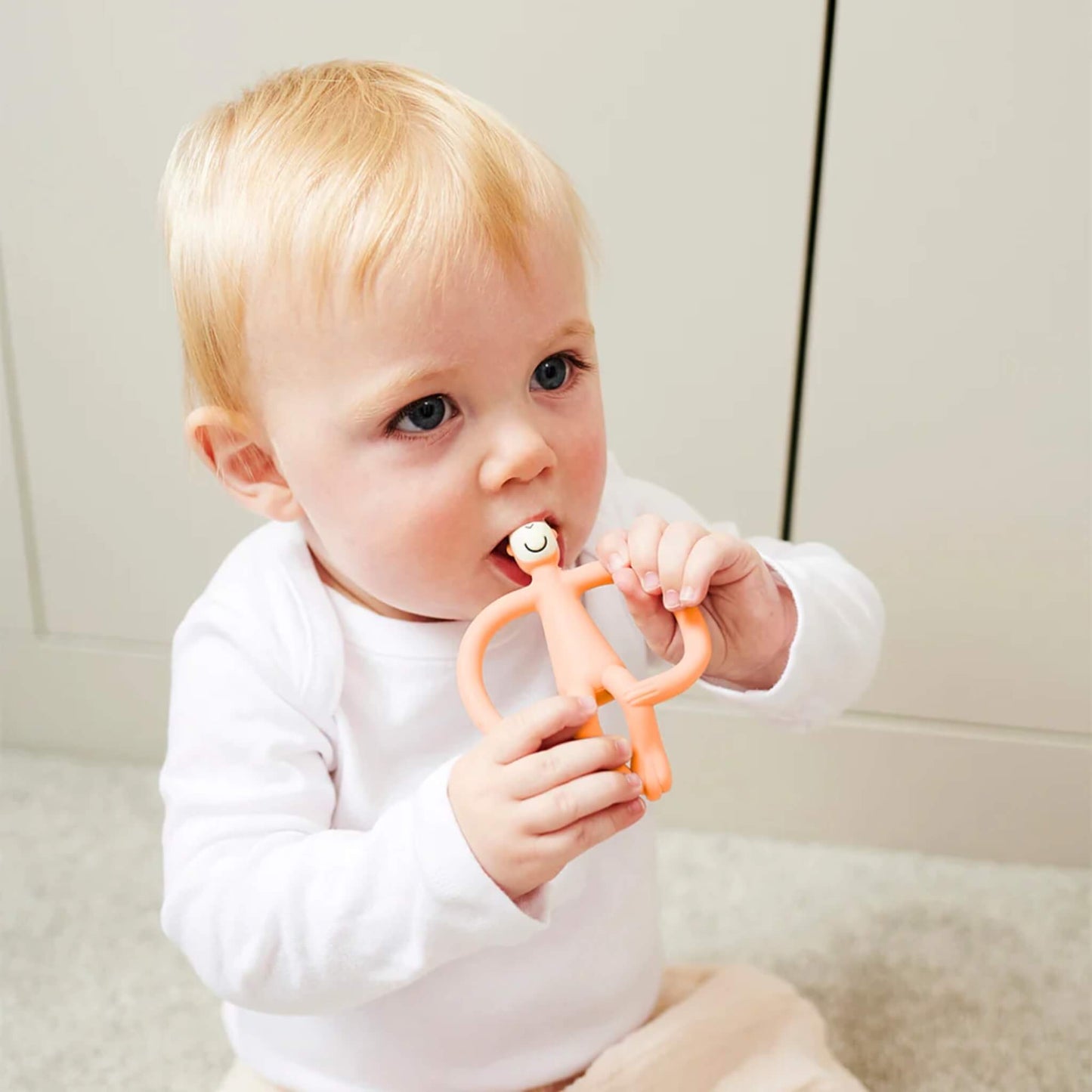 A baby sitting on a soft carpet holding a peach coloured monkey-shaped teething toy and chewing on the textured head.