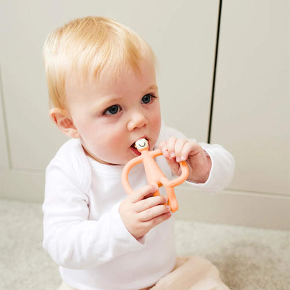 A baby sitting on a soft carpet holding a peach coloured monkey-shaped teething toy and chewing on the textured head.