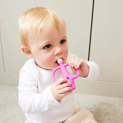 A baby sitting on a light-coloured floor holding a pink silicone teether with both hands and chewing on the smooth top section. The baby is wearing a white long-sleeved top and pale trousers.