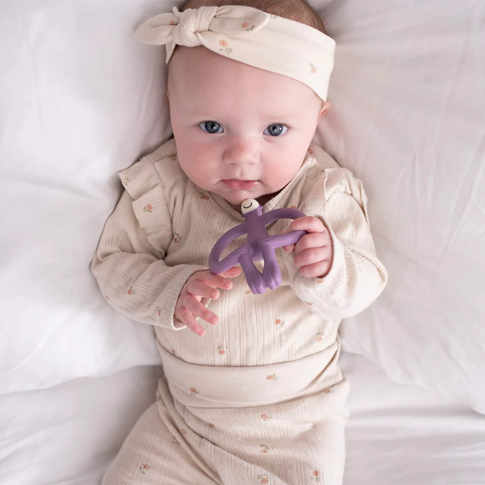 A baby lying on a white bed holding a purple monkey-shaped silicone teether while looking up toward the camera.