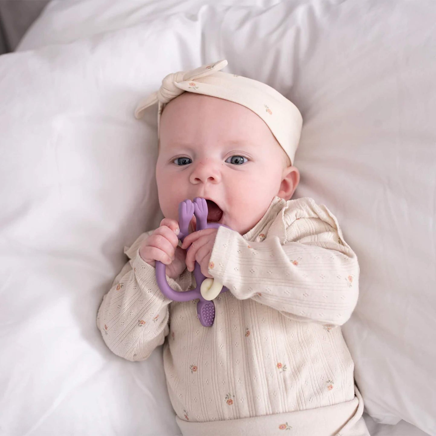 A baby lying on a white bed and chewing on a purple monkey-shaped silicone teether while holding it with both hands.