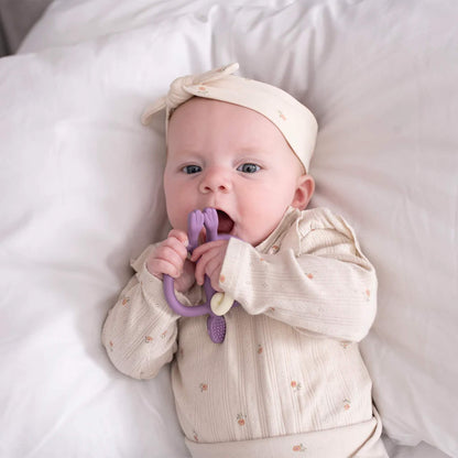A baby lying on a white bed and chewing on a purple monkey-shaped silicone teether while holding it with both hands.