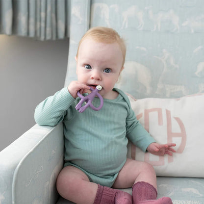 A baby sitting on a patterned nursery chair and mouthing a purple monkey-shaped silicone teether while wearing a mint-green ribbed bodysuit.
