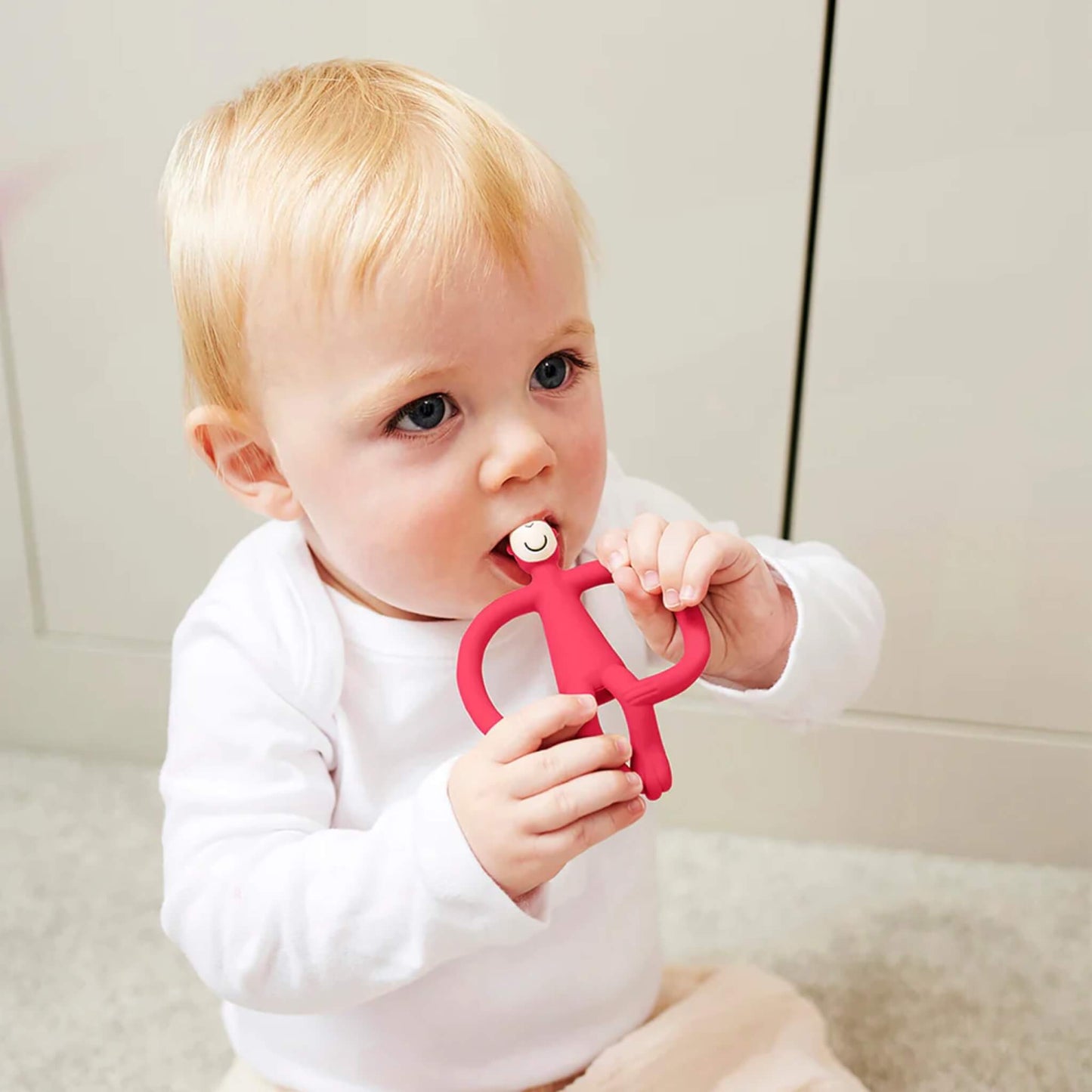 A baby sitting on a light-coloured floor holding a red silicone teether with both hands and chewing on the smooth top section. The baby is wearing a white long-sleeved top and pale trousers.