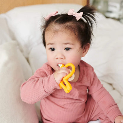 A baby sitting on a bed and chewing on a bright yellow silicone teether while wearing a rose-coloured romper.