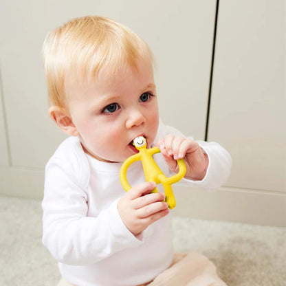 A baby sitting on a light-coloured floor holding a yellow silicone teether with both hands and chewing on the smooth top section. The baby is wearing a white long-sleeved top and pale trousers.