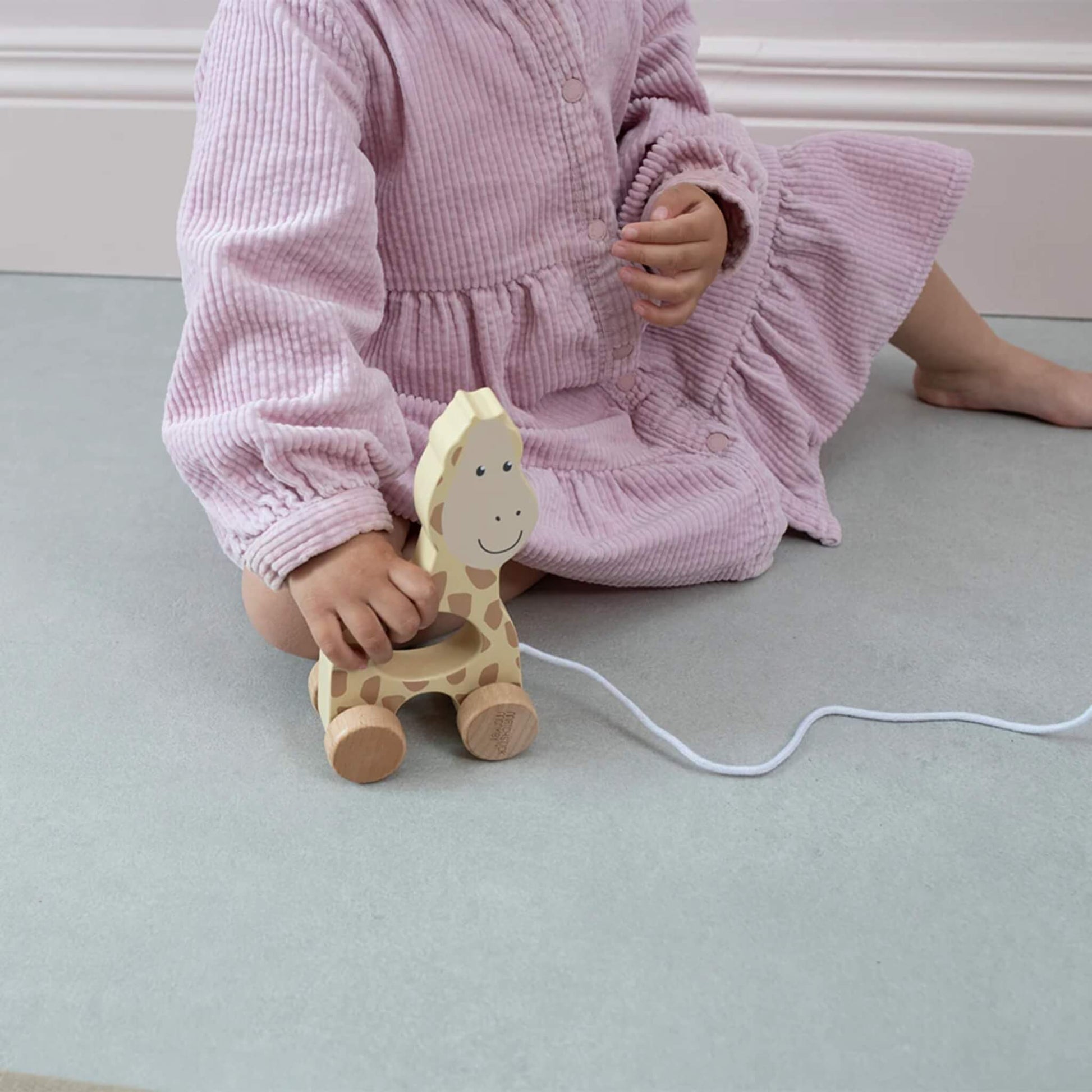 A toddler sitting on the floor wearing a pink corduroy outfit, holding a wooden giraffe pull toy by its cut-out centre.