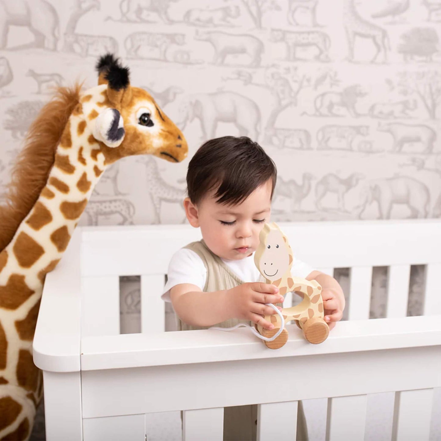 A young child standing at a white cot, holding a wooden giraffe pull toy while a large stuffed giraffe stands beside them.