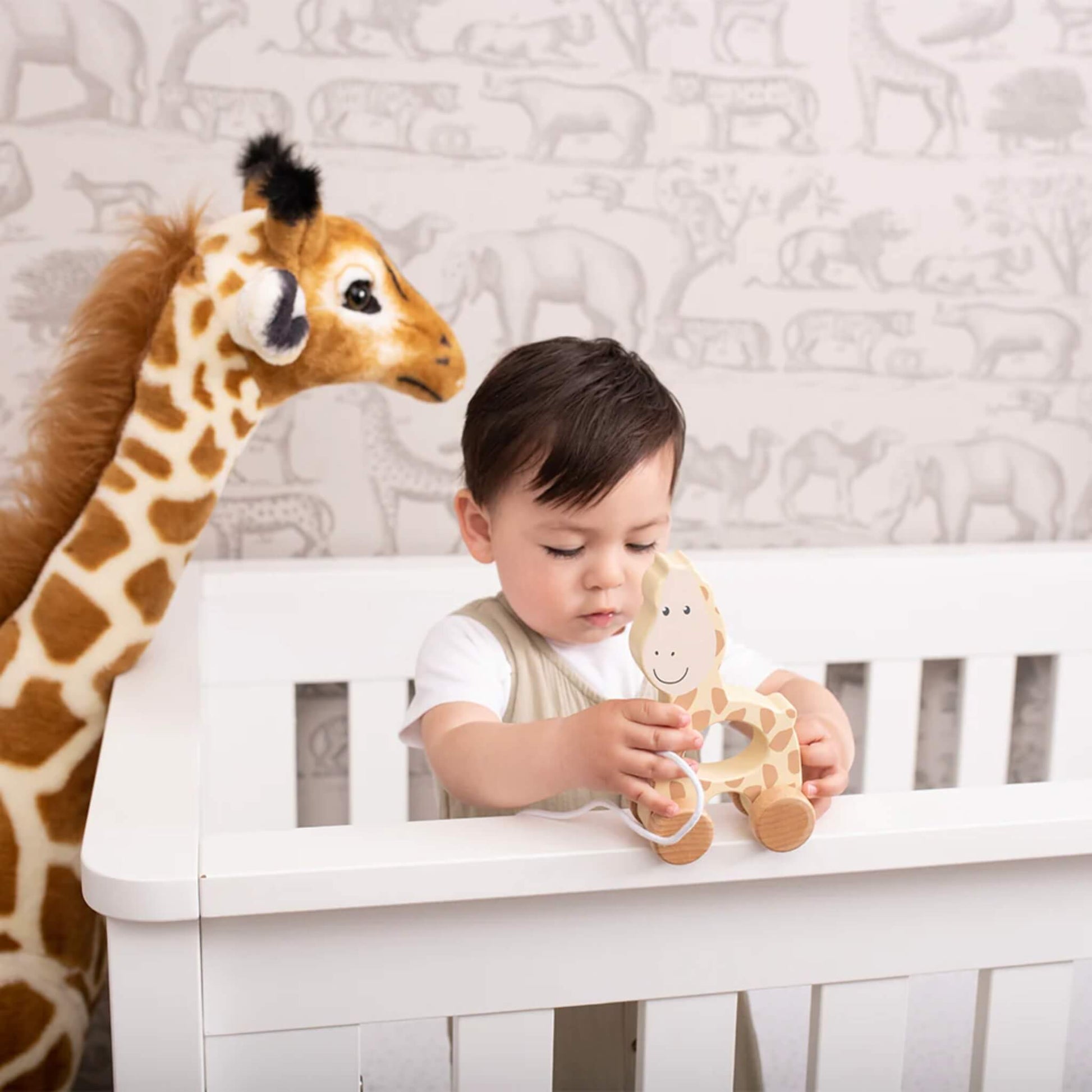 A young child standing at a white cot, holding a wooden giraffe pull toy while a large stuffed giraffe stands beside them.