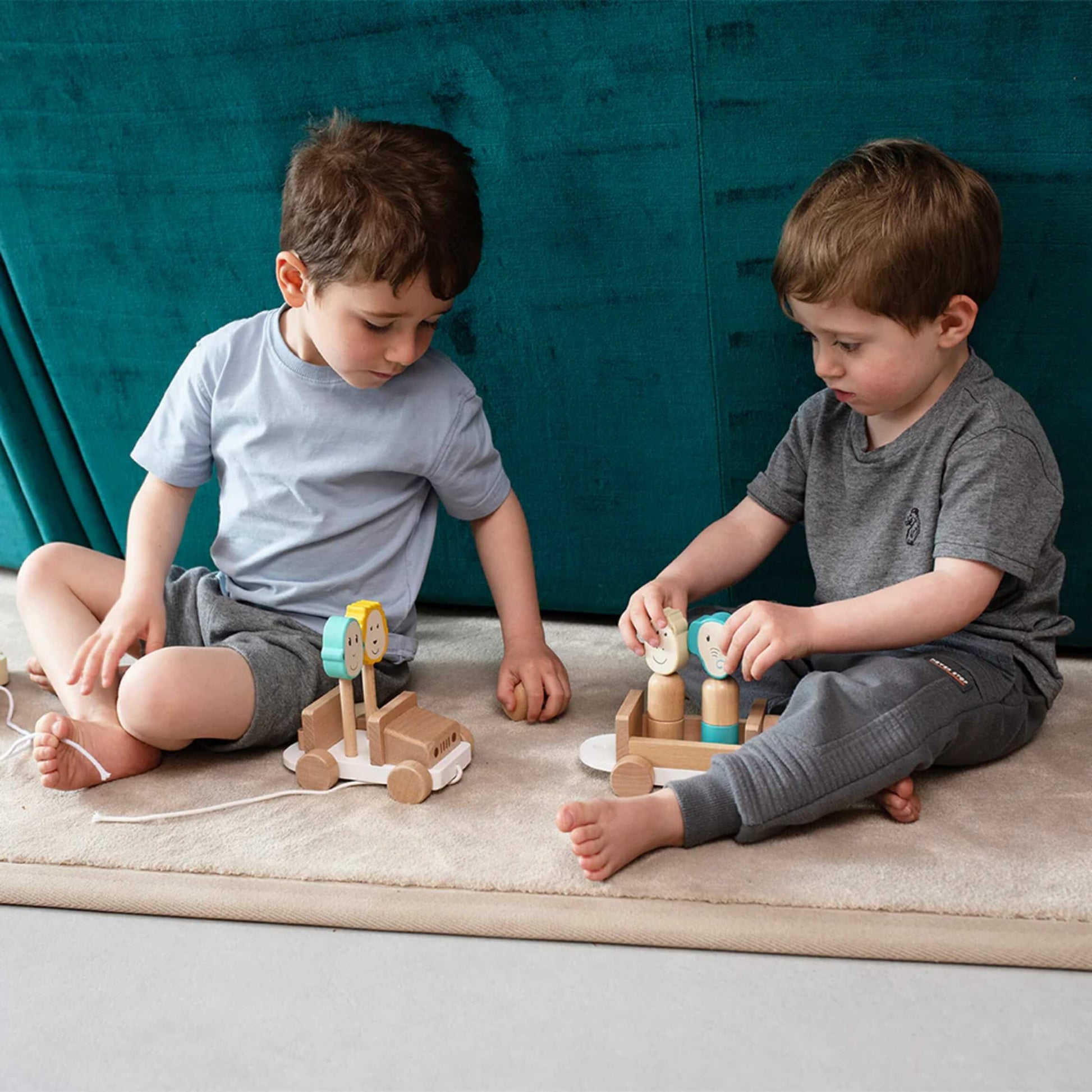 Two young children sitting on a soft rug, each playing with the wooden safari jeep toy and placing the animal pieces into the seats.