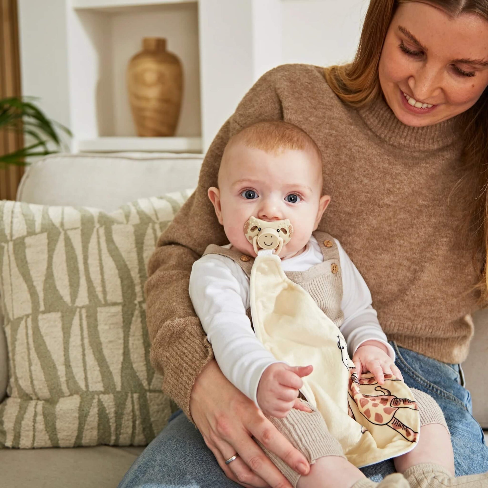 Baby sitting on an adult’s lap using the giraffe-themed silicone soother while holding the attached cream comforter.