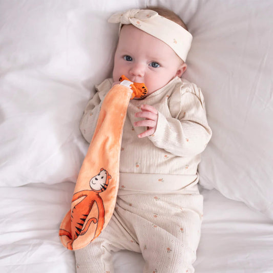 A baby lying on a bed holding the orange comforter while chewing on the attached tiger-face silicone soother.