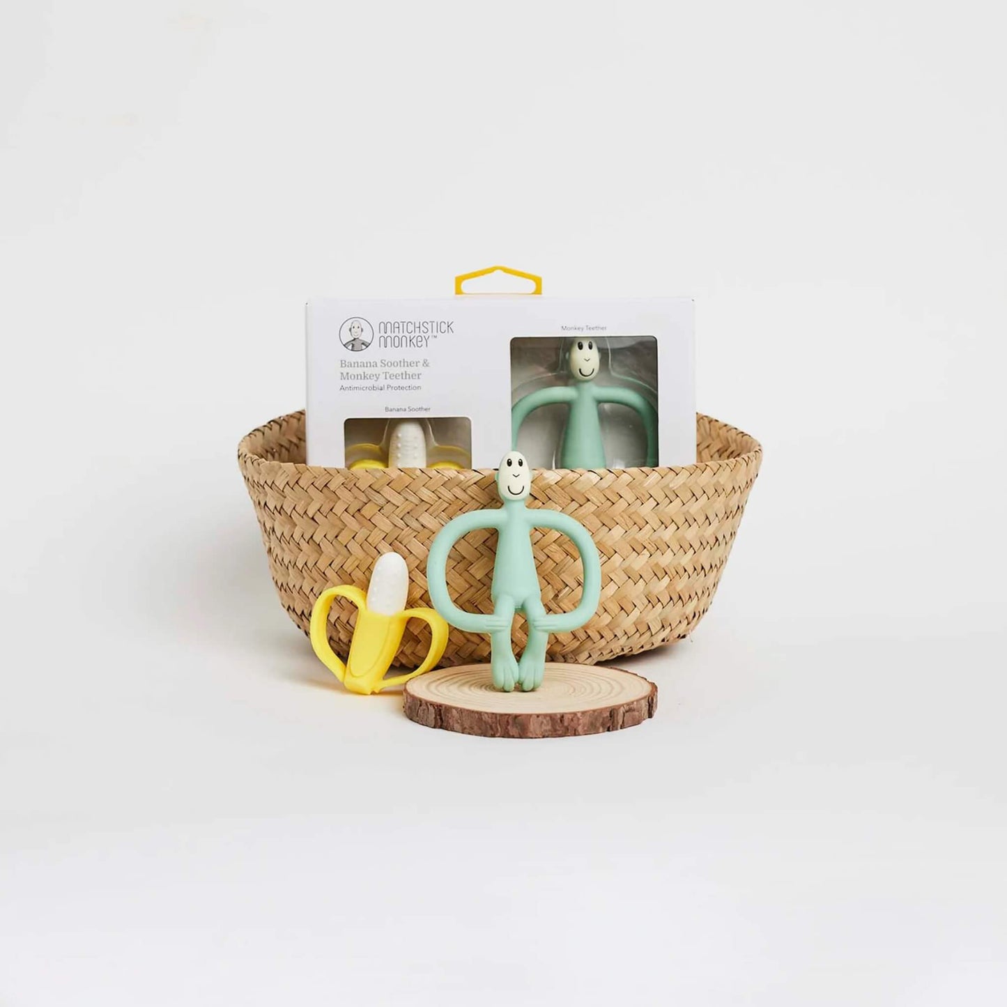 A woven basket on a white surface containing the product box, with the banana soother and monkey teether displayed in front on a wooden slice.