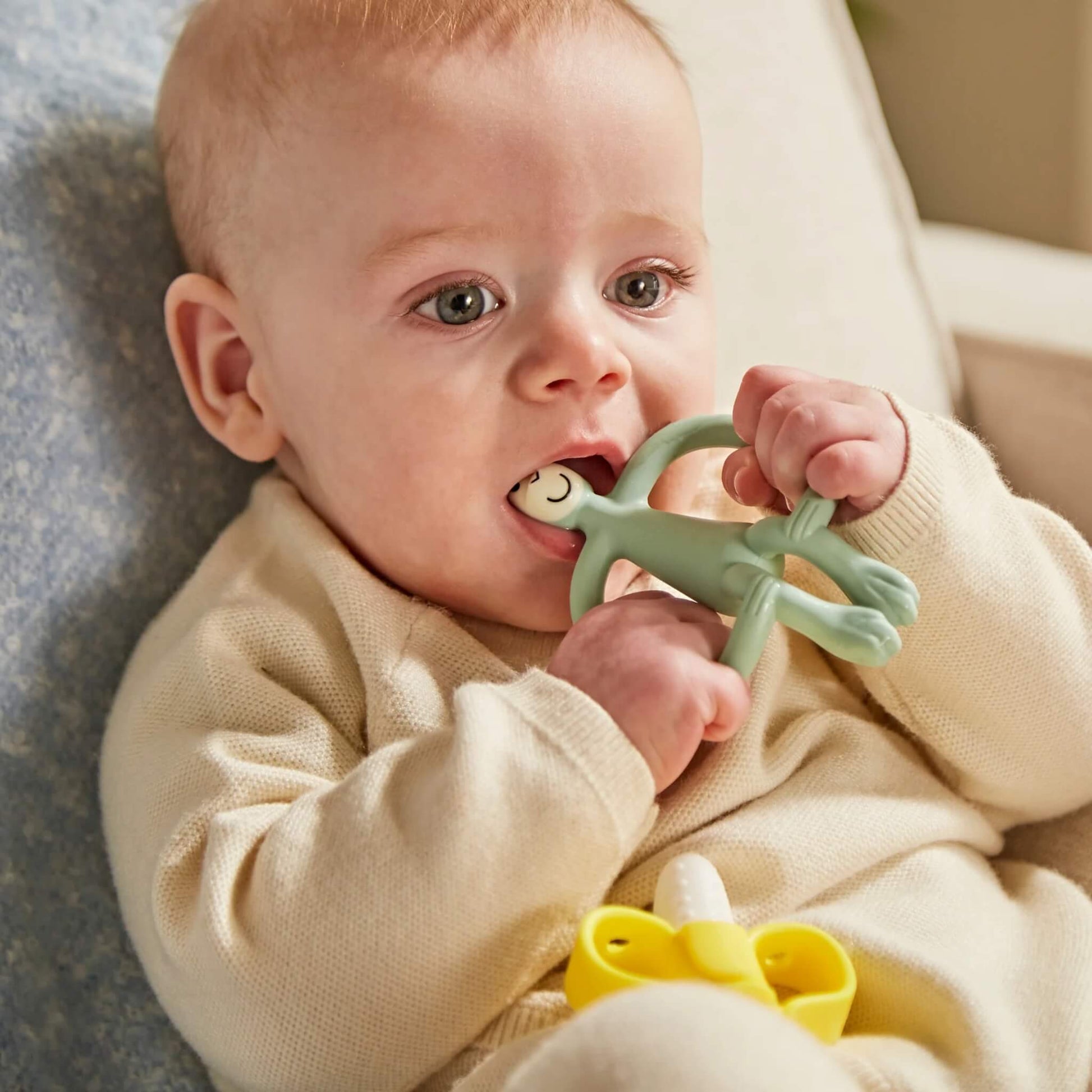 A baby chewing on a sage green monkey-shaped silicone teether while sitting on a soft chair, holding the teether with both hands.