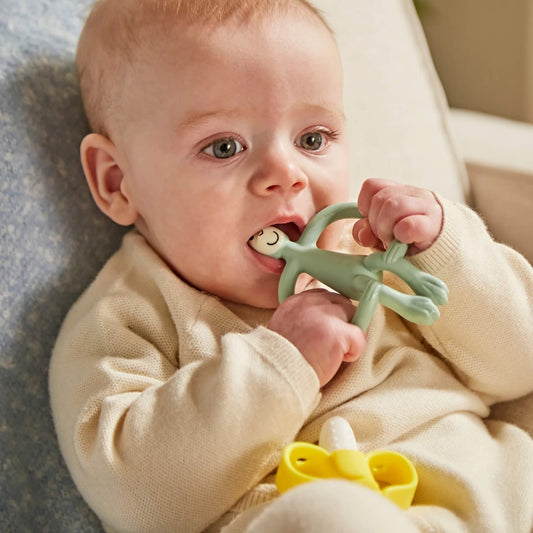 A baby chewing on a sage green monkey-shaped silicone teether while sitting on a soft chair, holding the teether with both hands.
