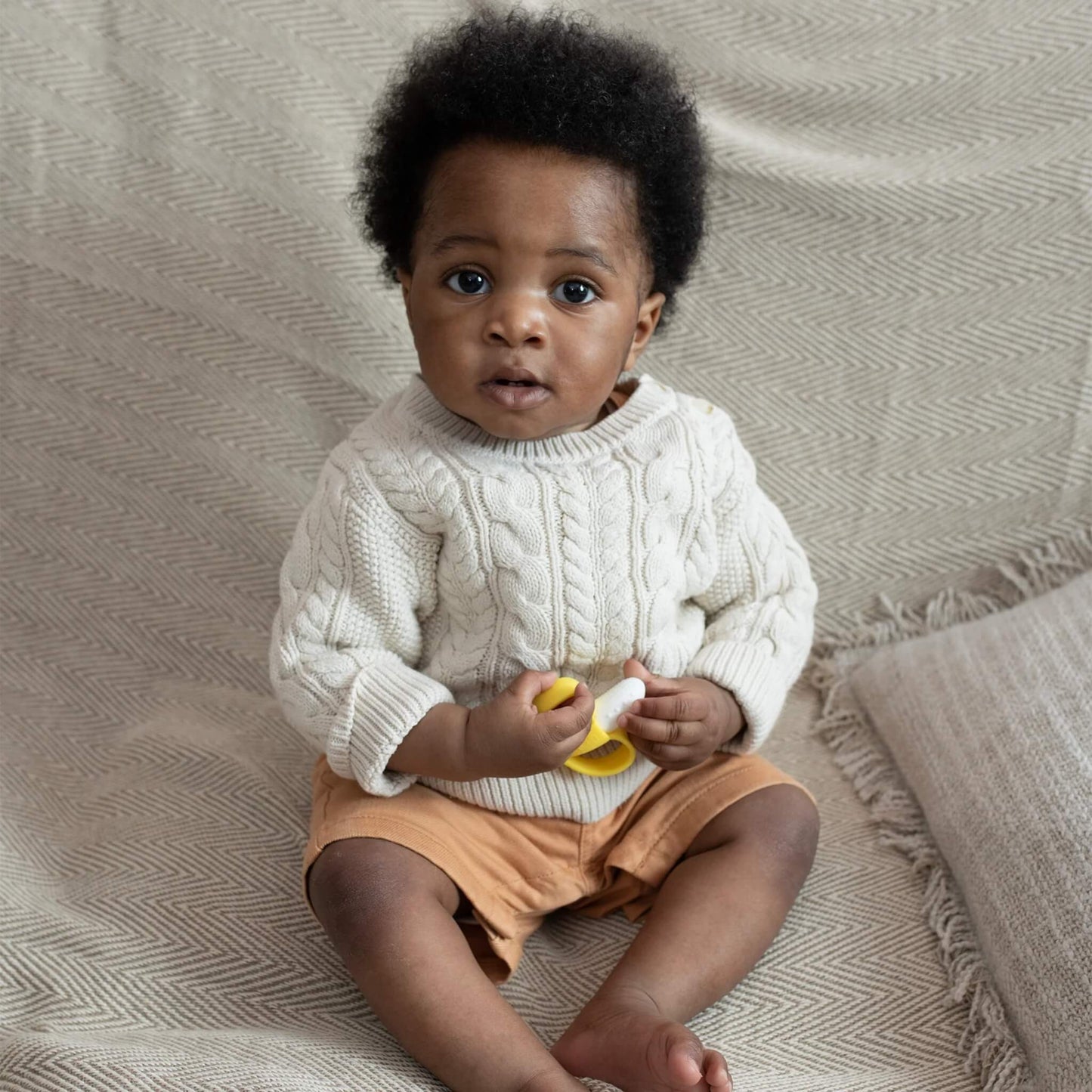 A baby sitting on a textured blanket holding a yellow and white banana-shaped silicone soother in both hands.