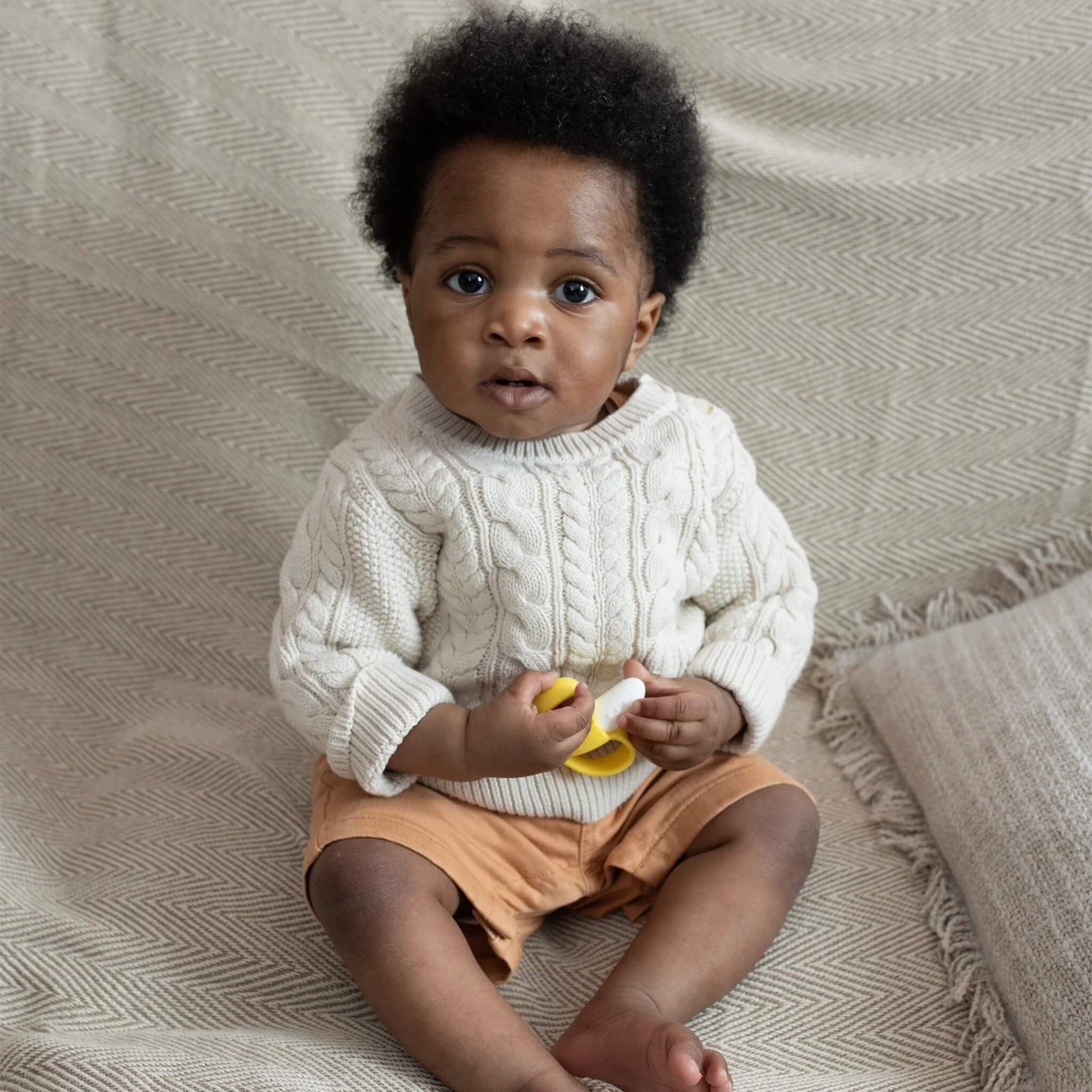 A baby sitting on a textured blanket holding a yellow and white banana-shaped silicone soother in both hands.