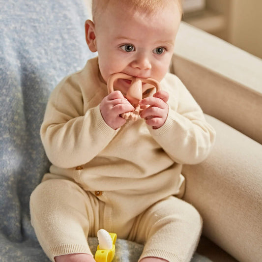 A baby wearing a cream knitted outfit sitting on a sofa and chewing a dusty pink silicone teether with both hands.