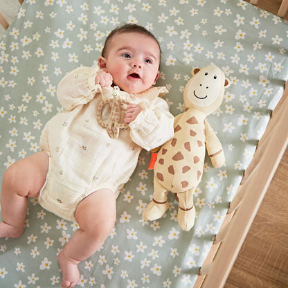 A baby lying on a daisy-patterned cot sheet holding a giraffe-shaped teething ring beside a soft giraffe comfort toy.