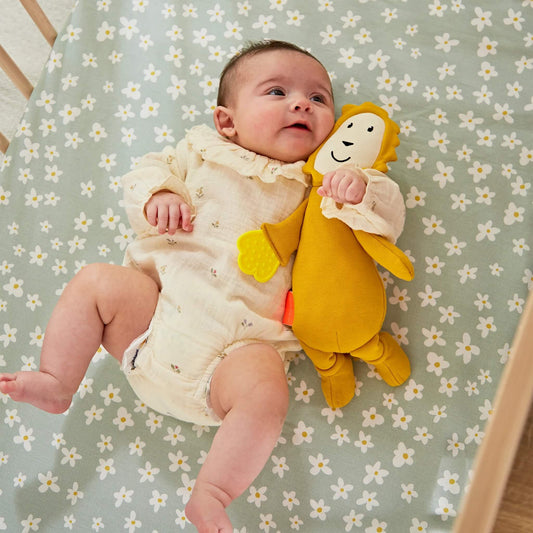 A baby lying on a daisy-patterned sheet cuddling a soft lion sensory toy with an attached textured teething leaf.