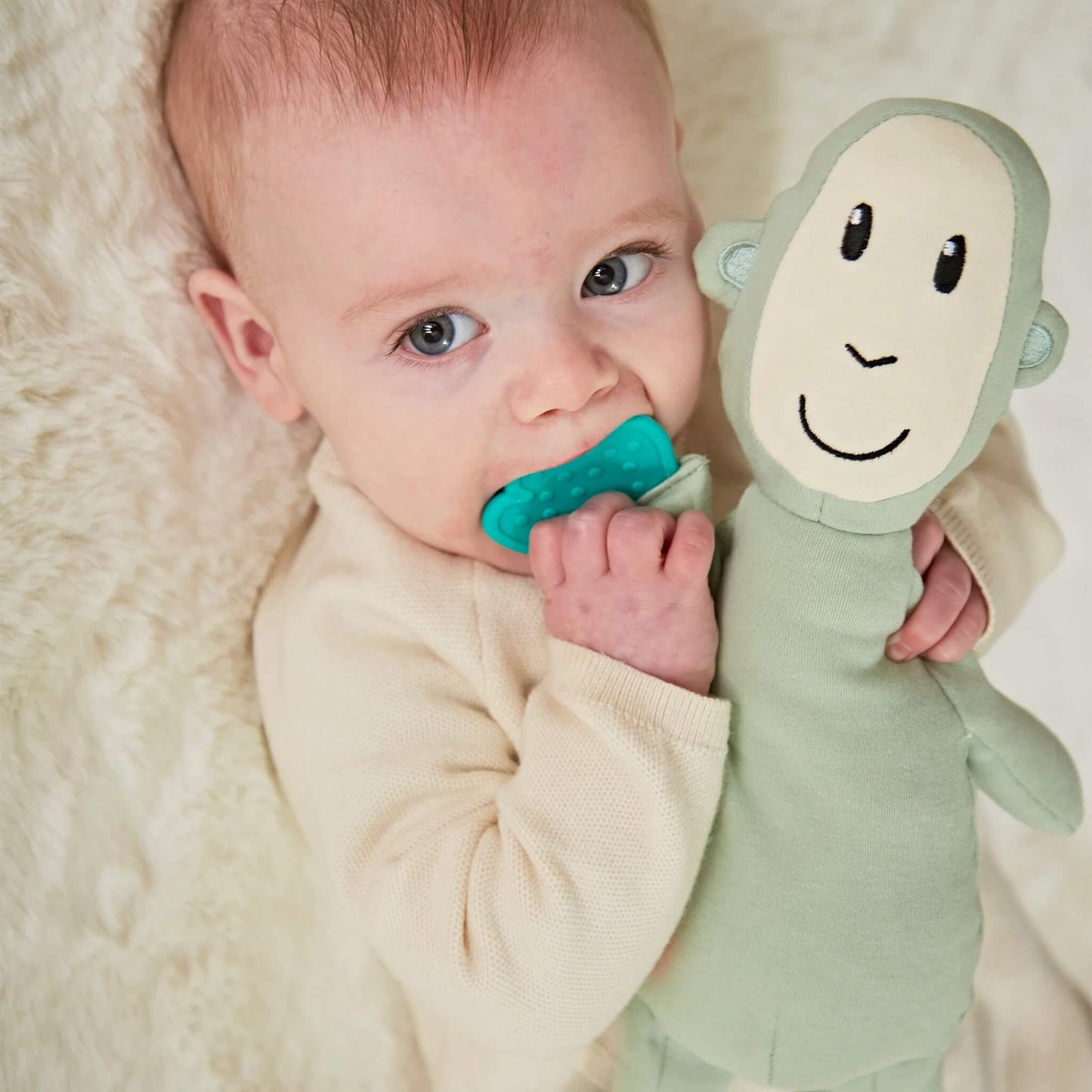 A baby lying on a cream blanket chewing a textured teething leaf while cuddling a soft green monkey sensory toy.