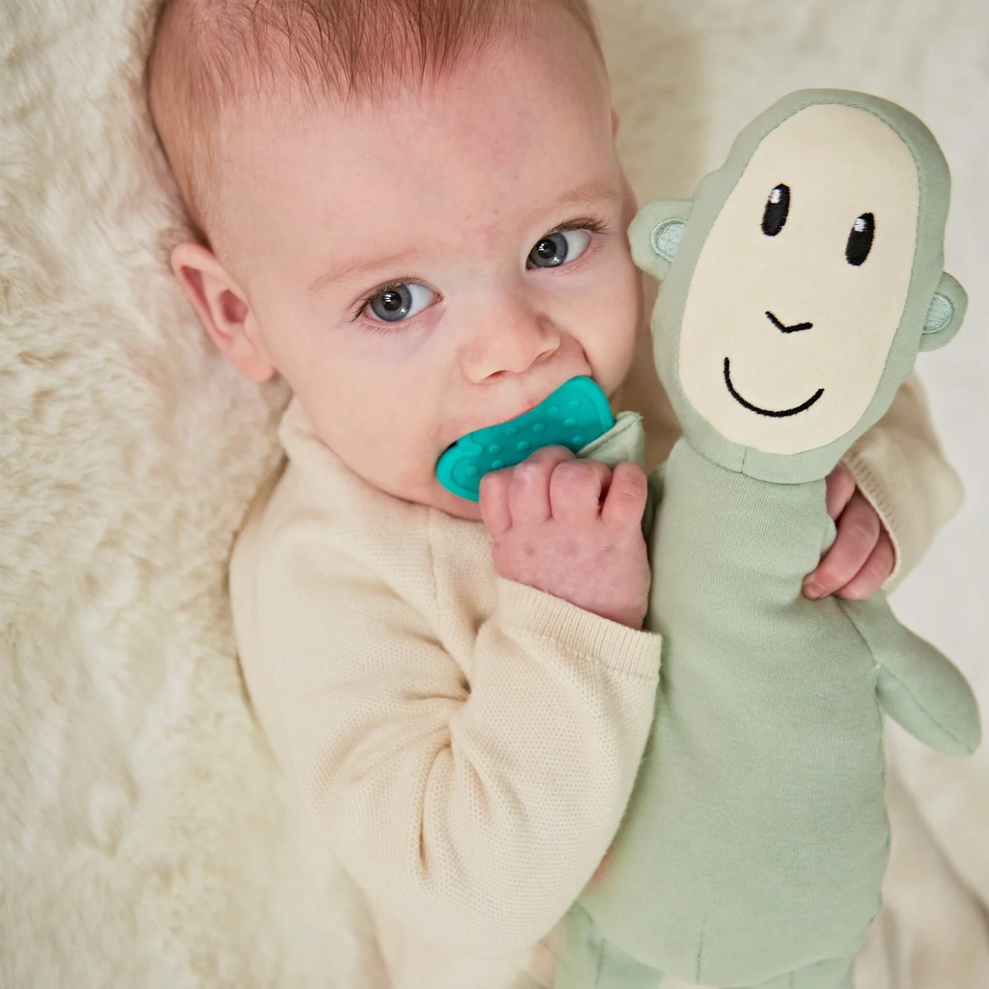 A baby lying on a cream blanket chewing a textured teething leaf while cuddling a soft green monkey sensory toy.