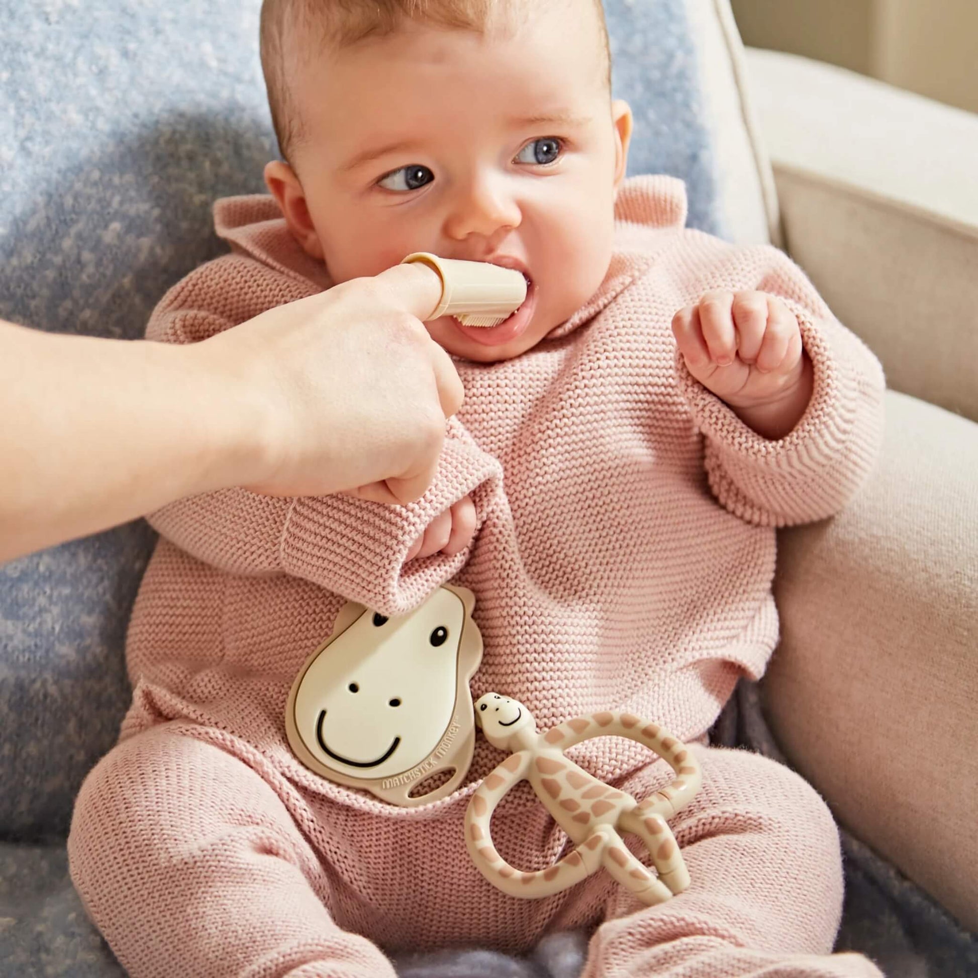 A baby sitting on a chair while an adult gently brushes the baby's gums with a silicone finger toothbrush.