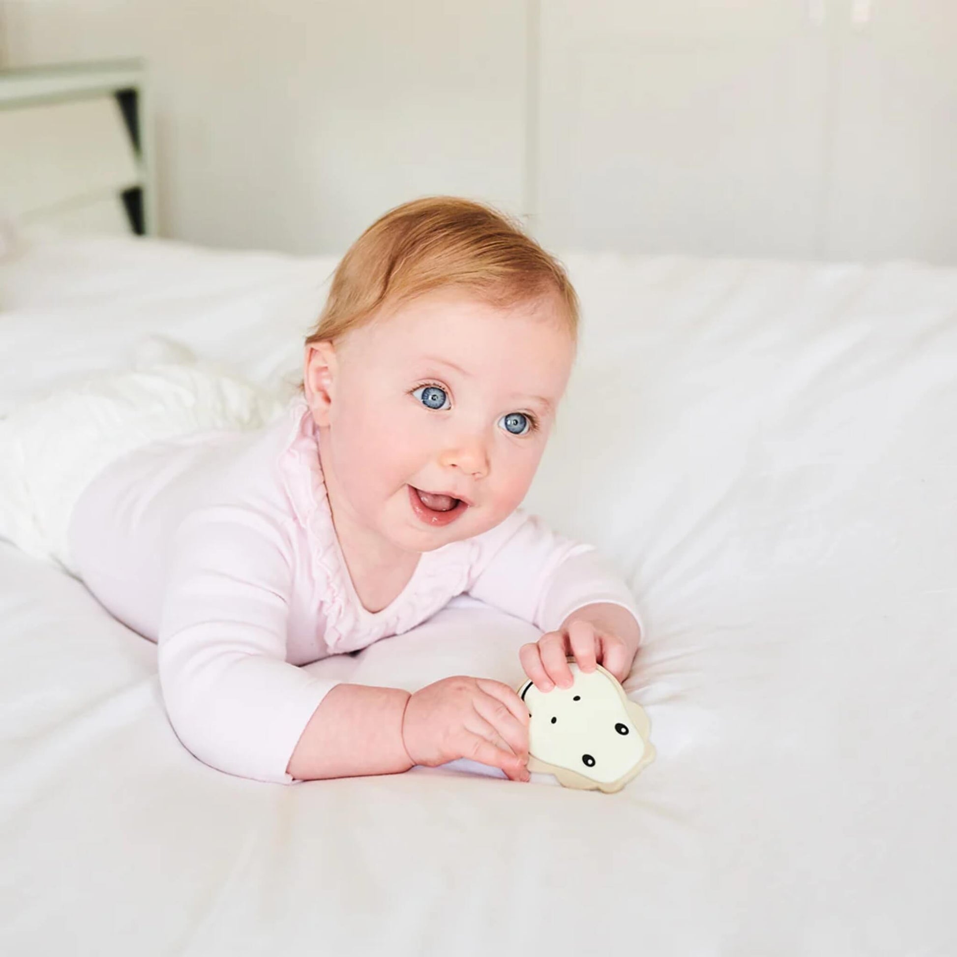 A baby lying on a white bed holding a flat giraffe-shaped teether in both hands.