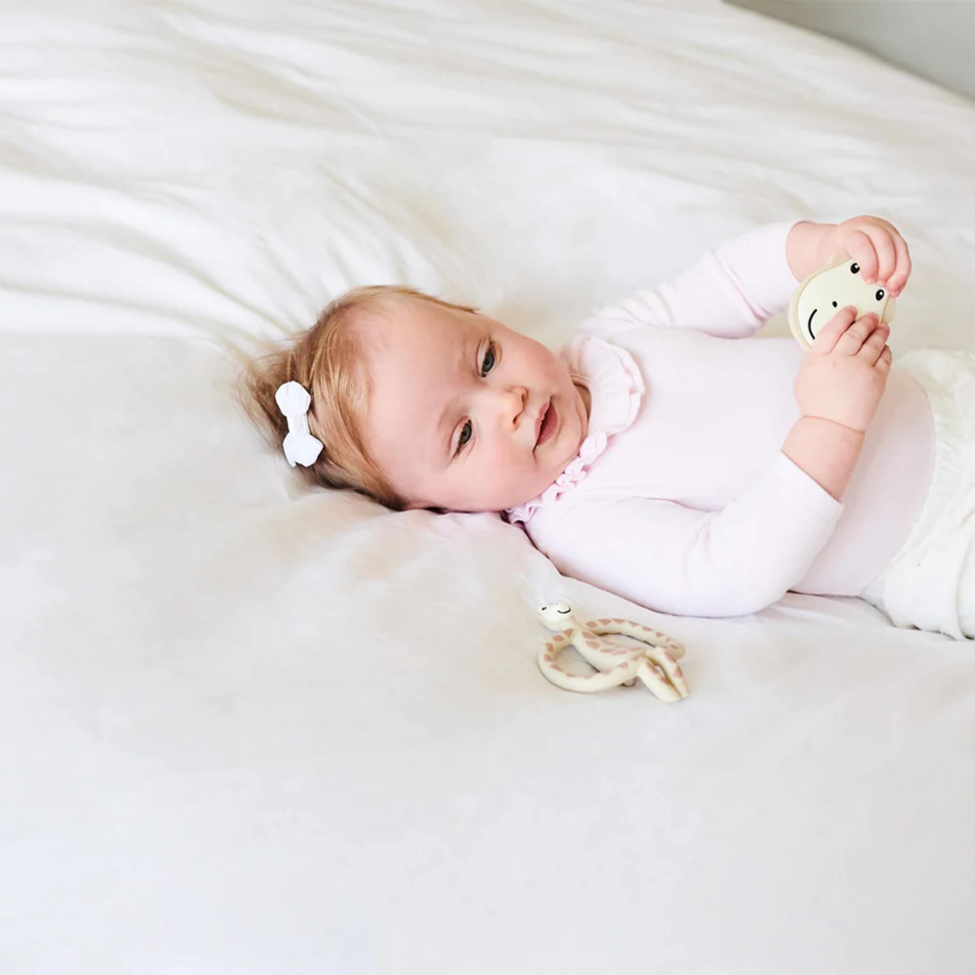 A baby lying on a bed holding a flat teether, with a giraffe-shaped teether resting beside them.