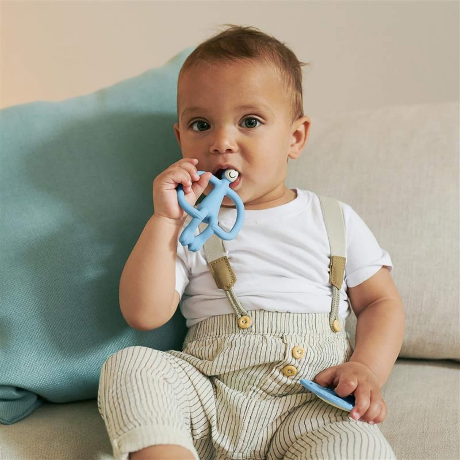 A toddler sitting on a sofa holds a flat teether and chews a blue animal-shaped silicone teether.