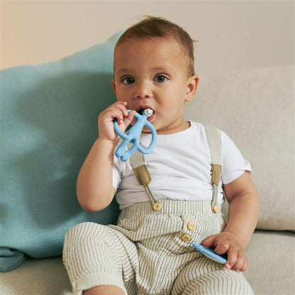 A toddler sitting on a sofa holds a flat teether and chews a blue animal-shaped silicone teether.