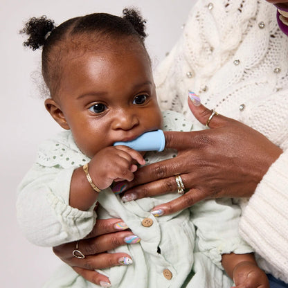 A baby in a pale green outfit chews a blue silicone finger toothbrush while being held by a woman.