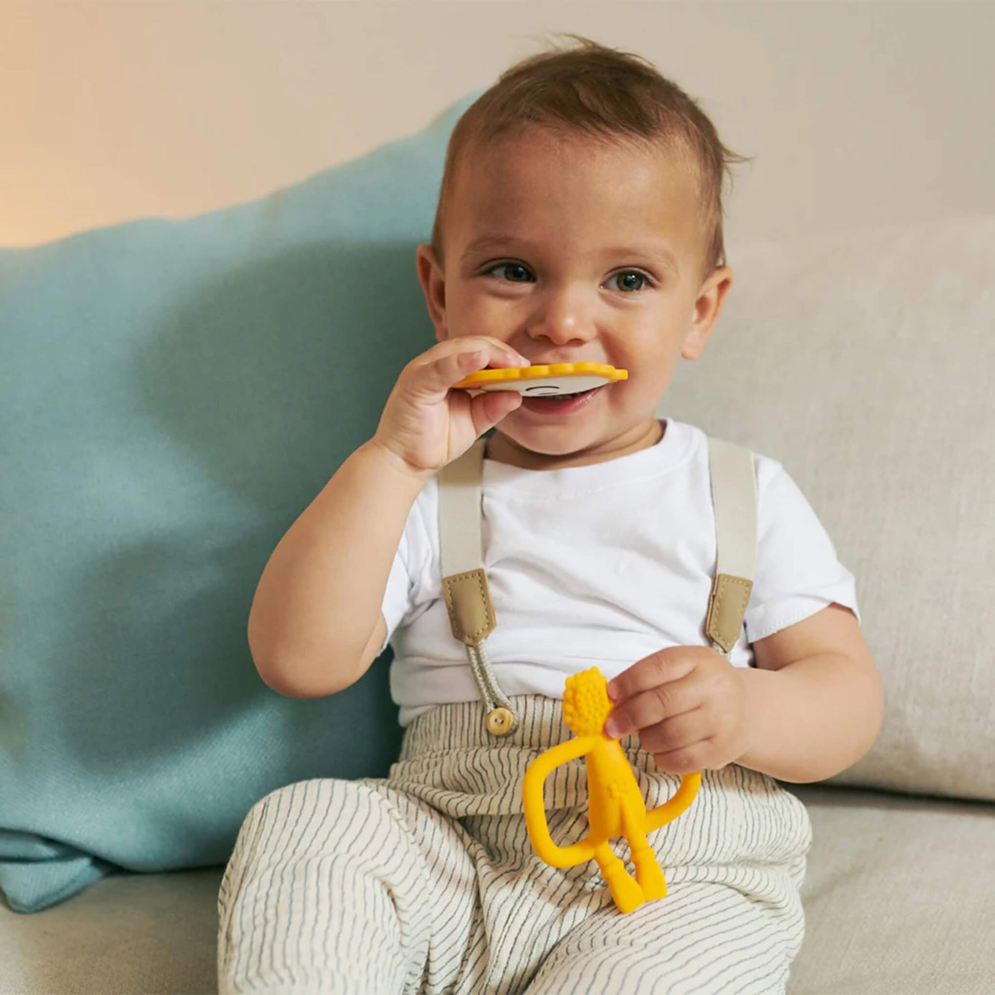 A toddler sitting on a sofa holding a flat lion-shaped teether and lifting it towards their mouth.
