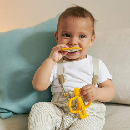 A toddler sitting on a sofa holding a flat lion-shaped teether and lifting it towards their mouth.