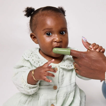 A baby in a pale green dress chews an olive silicone finger toothbrush while a woman gently supports them.