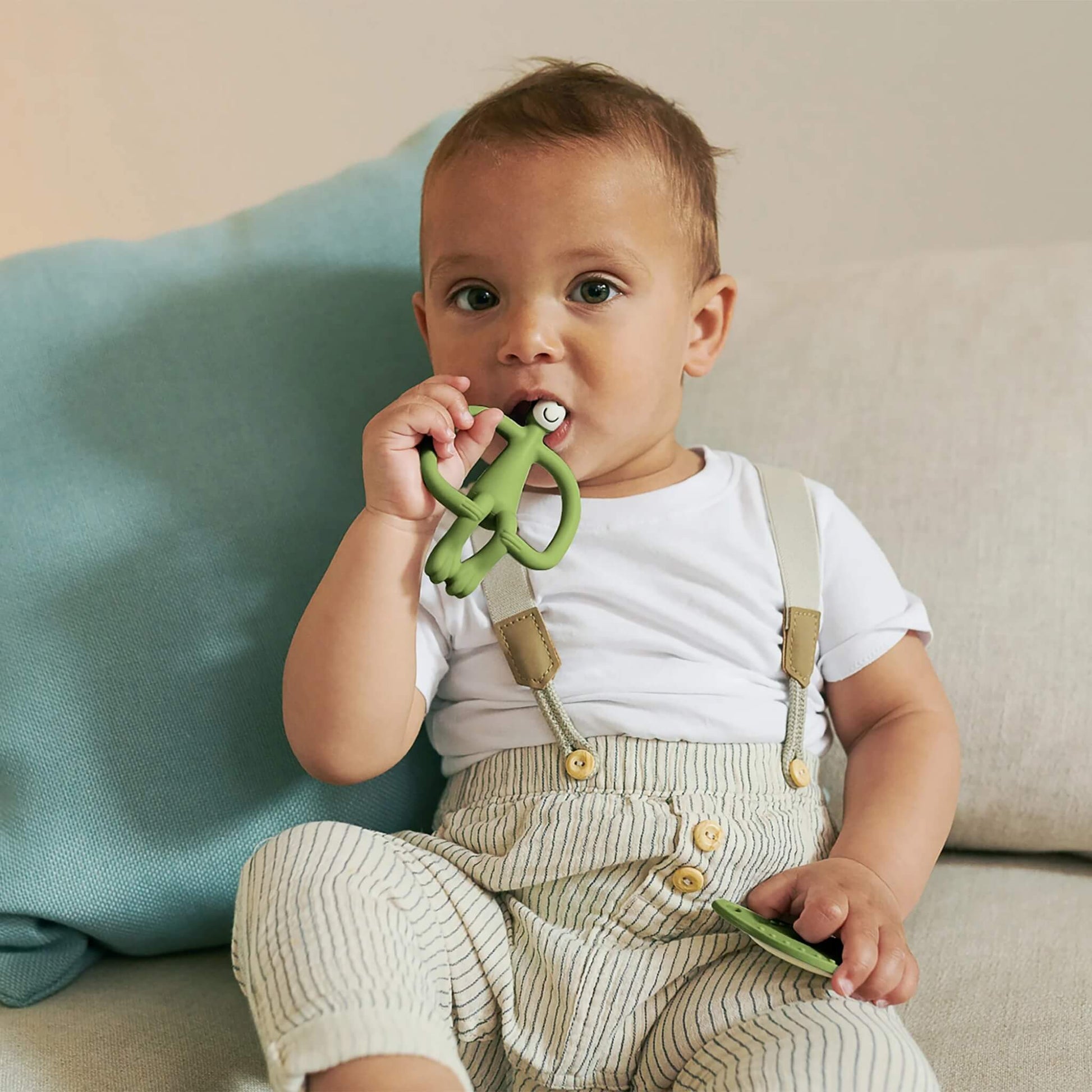 A toddler sitting on a sofa chews an olive animal-shaped silicone teether while holding a matching flat teether.