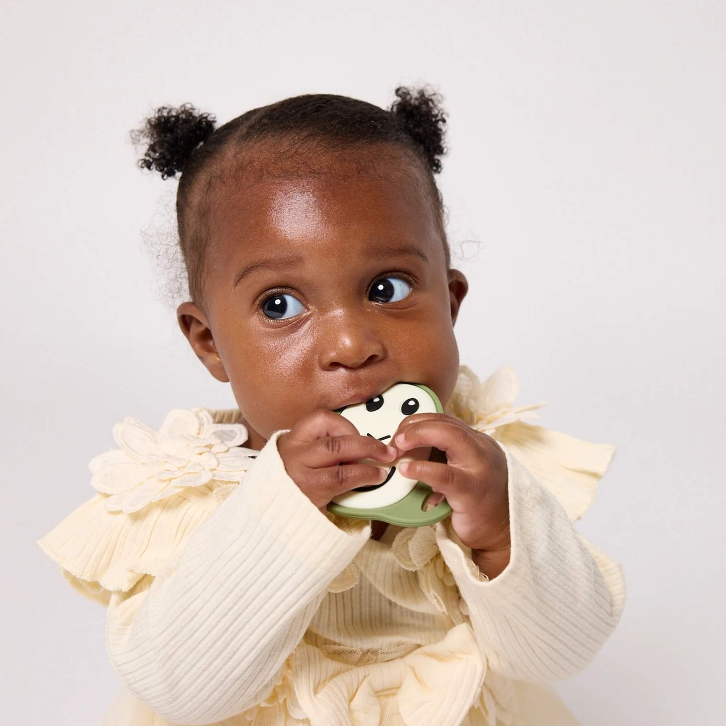 A baby wearing a cream outfit holds and chews an olive flat silicone teether with a smiling face design.
